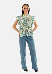 Bluse mit Blumenmuster und Rüschensleeves in grünen und blauen Farbtönen, kombiniert mit hellblauen, weiter geschnittenen Jeans und schwarzen High Heels.