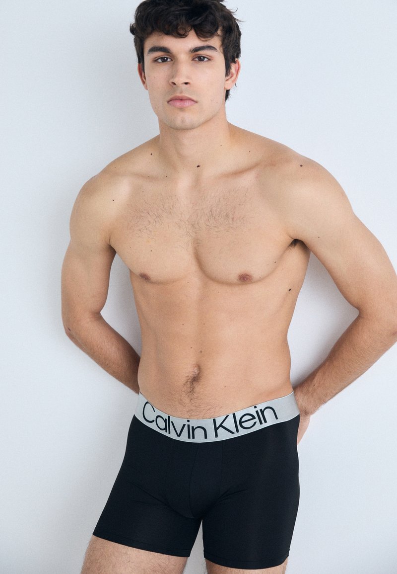 Boxers noirs avec une ceinture grise portant "Calvin Klein" en noir. Tissu lisse avec un design ajusté.
