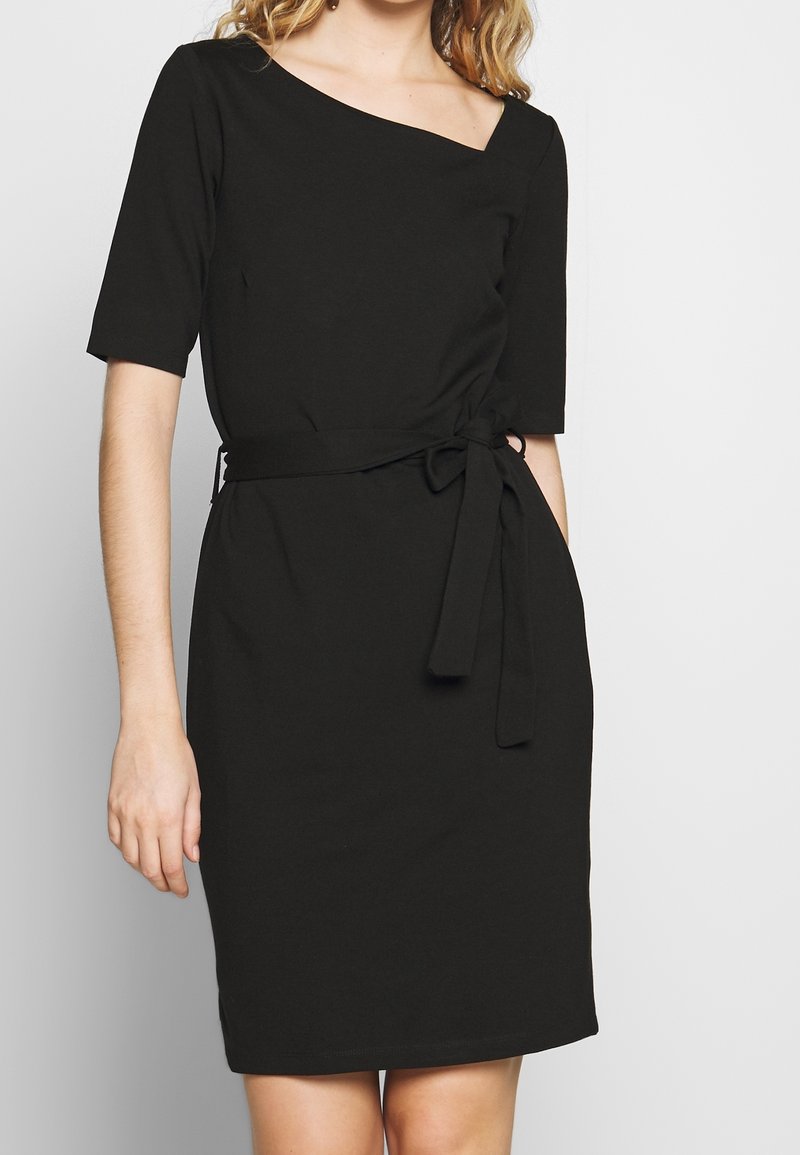 Femme portant une robe noire mi-longue avec des manches au niveau des coudes, un décolleté asymétrique et une ceinture en tissu nouée à la taille.