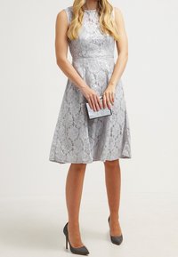 Kvinna i ärmlös silverfärgad spetsklänning som håller en liten glittrig clutch, bär grå klackskor, stående mot en enfärgad bakgrund.