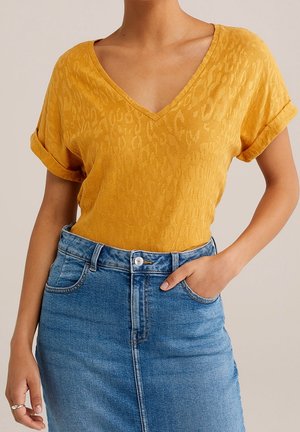 Kobieta ubrana w musztardowożółty, teksturowany top z krótkim rękawem i dekoltem w serek, włożony do wysokiego stanem niebieskiej spódnicy dżinsowej, z jedną ręką w kieszeni.