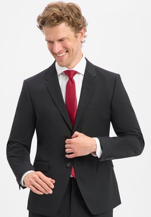 Mann lächelt, während er eine schwarze Anzugjacke über einem weißen Hemd und einer roten Krawatte vor einem einfarbigen Hintergrund zuknöpft.