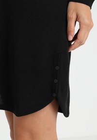 Abito nero lavorato a maglia con orlo arrotondato, caratterizzato da quattro bottoni neri lungo il lato, che mette in risalto una texture liscia e un design aderente.