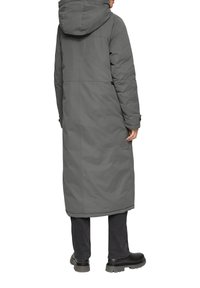 Long manteau rembourré gris avec capuche, présentant une coupe droite et un léger éclat. Les détails incluent des poches et des poignets réglables, associés à un pantalon sombre et des bottes noires.