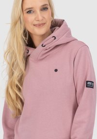 Donna bionda che sorride, indossando una felpa rosa polveroso con occhiello per cordini neri e un patch sul braccio sinistro.