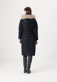 Giacca puffa blu navy con cappuccio foderato in pelliccia ecologica, texture a righe verticali, design lungo fino al ginocchio e maniche lunghe, indossata sopra pantaloni grigi e stivali neri.