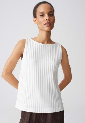 Femme aux cheveux courts et bouclés, portant un haut blanc sans manches côtelé et un pantalon sombre, debout les mains dans le dos.