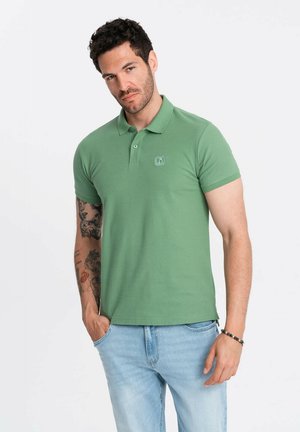 Camisa polo verde de algodón, con cuello y mangas cortas. Incluye un pequeño logo en el lado izquierdo del pecho. Llevada con jeans de color azul claro.
