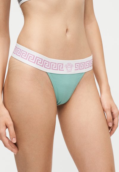 Femme portant un string côtelé vert avec une ceinture blanche ornée d'un motif clé grecque rose et d'un logo tête de Méduse au centre.