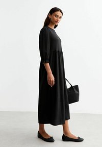 Black, long-sleeve dress with a textured finish and fitted bodice, flowing to an A-line skirt. Paired with black ballet flats and a bag.