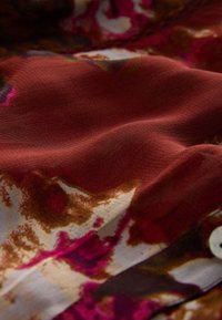 Tessuto morbido con una texture ondulata, caratterizzato da una combinazione di tonalità rosso scuro, rosa e marrone, con motivi floreali e un dettaglio di bottone bianco.