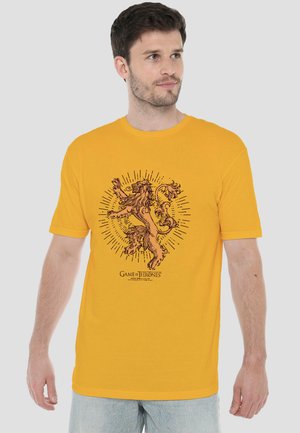 Mann trägt gelbes T-Shirt mit dunklem Löwenemblem und dem Schriftzug „Game of Thrones“, steht vor einem schlichten grauen Hintergrund.