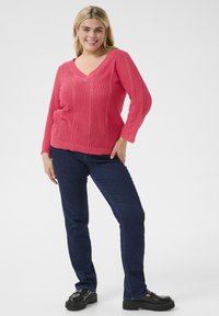 Pull en tricot corail avec un col en V, motif texturé et poignets côtelés, associé à un jean bleu foncé et des mocassins à plateforme noirs. Look lumineux et décontracté.