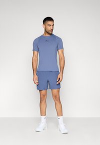 Μπλε αθλητική μπλούζα με κοντά μανίκια, που φέρει μαύρο λογότυπο, συνδυασμένη με αντίστοιχα μπλε σορτς. Φοριέται με λευκά αθλητικά παπούτσια και κάλτσες.