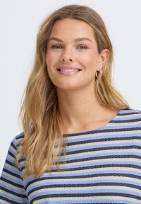 Mujer sonriente con cabello largo y rubio, que lleva pendientes de aro dorados y una blusa de rayas azules, negras y blancas sobre un fondo liso.