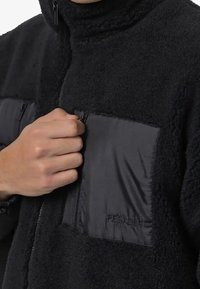 Ręka zapinająca czarną polarową kurtkę z zamkiem błyskawicznym, posiadającą kieszeń na piersi z zamkiem i haftowanym napisem "PEGADOR."