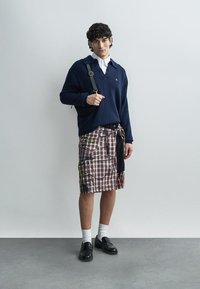 Pull en maille bleu marine, chemise blanche à col, jupe à carreaux avec poches, accents noirs, associée à des chaussettes blanches et des chaussures noires. Fond minimaliste.