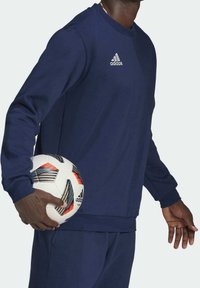 Sweatshirt Adidas bleu marine à manches longues et col rond, avec un petit logo sur la poitrine. Un ballon de football est tenu à la taille.