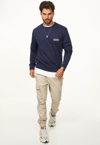 Navy sweatshirt with white graphic detail, worn over a white T-shirt, paired with beige cargo pants and white sneakers, on a light background.