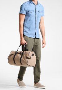 Hombre vestido con una camisa de botones de manga corta azul y pantalones oliva, sosteniendo una bolsa de deporte beige con correas negras, de pie sobre un fondo blanco.