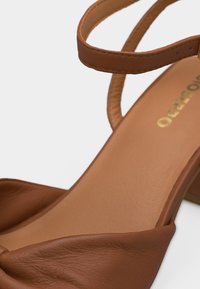 Καφέ δερμάτινο σανδάλι με λεπτό λουράκι στον αστράγαλο και επενδυμένη διπλωμένη λεπτομέρεια στο μπροστινό μέρος σε φωτεινό φόντο.