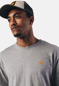 Šedé bavlněné tričko s kulatým výstřihem a malým oranžovým emblémem slona na levé straně hrudi. Texturovaná tkanina s jemným povrchem.