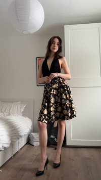 Jonge vrouw in een zwarte haltertop en een zwarte-gouden bloemenrok staand in een moderne slaapkamer met houten vloer en een gitaar achter haar.