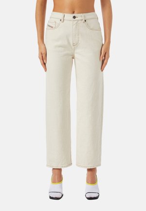 Femme portant un jean crème taille haute à jambes droites et des chaussures blanches ouvertes à bout ouvert avec des semelles noires et un liseré jaune.