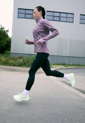 Lila futódzseki bélelt textúrával, fekete leggings perforált díszítéssel, és világoszöld futócipő. Fehér zoknik láthatóak.