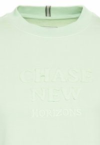 Jasnozielona bluza z ribbowym kołnierzem, ozdobiona haftowanym napisem "CHASE NEW HORIZONS" w kontrastowej fakturze i kolorze.