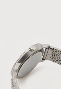 Ασημένιο ρολόι χειρός από ανοξείδωτο ατσάλι με πλεκτό μπρασελέ, σε γωνία που δείχνει το κορώνα με το λογότυπο "CK" και την πίσω θήκη, πάνω σε απλό λευκό φόντο.