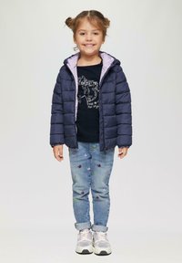 Marineblauwe pufferjack met een paarse voering, zwart T-shirt met grafische print, lichtblauwe jeans met kleurrijke vlinderborduursels en witte sneakers.