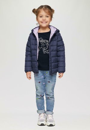 Granatowa kurtka pikowana z fioletowym podbiciem, czarny t-shirt z grafiką, jasnoniebieskie dżinsy z kolorową haftowaną motylkową aplikacją oraz białe sneakersy.
