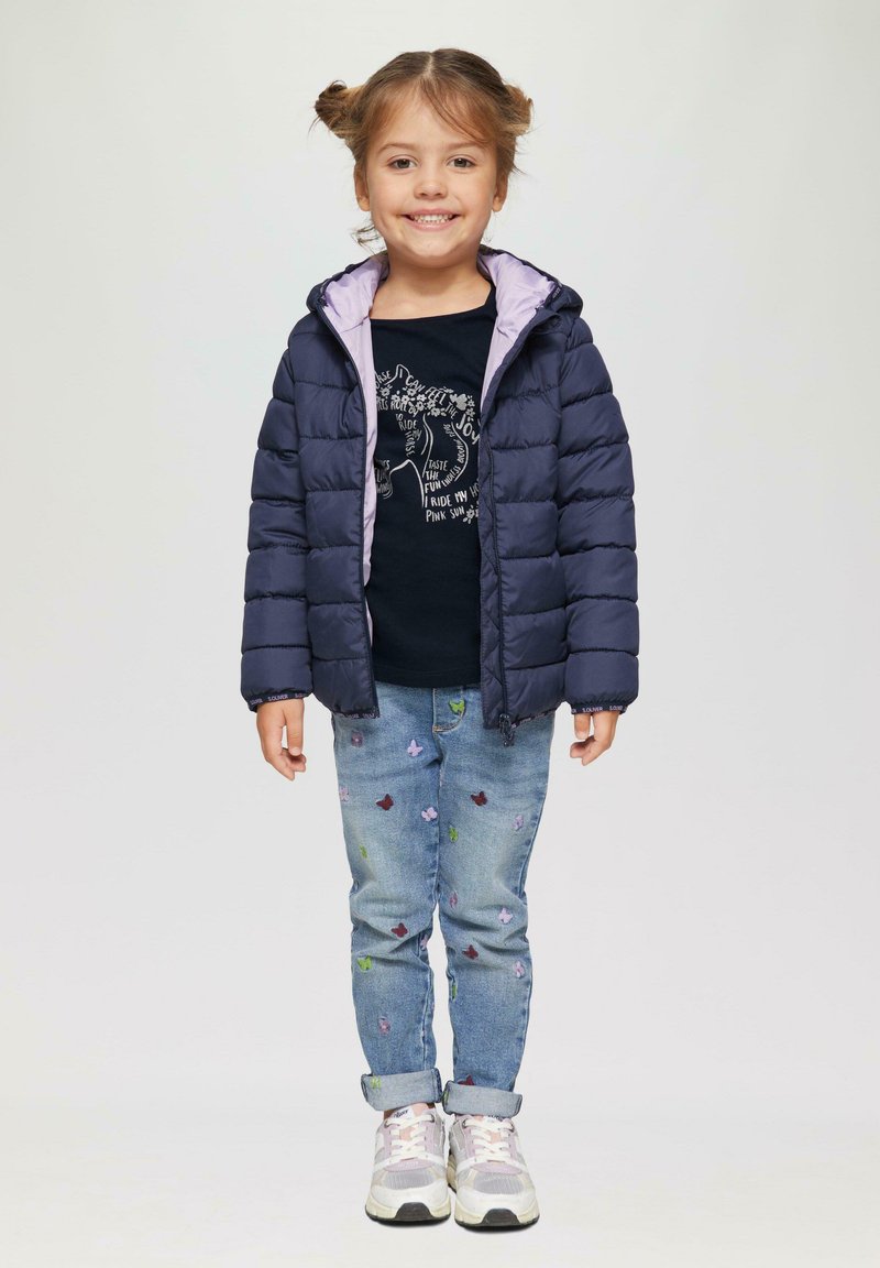 Marineblauwe pufferjack met een paarse voering, zwart T-shirt met grafische print, lichtblauwe jeans met kleurrijke vlinderborduursels en witte sneakers.