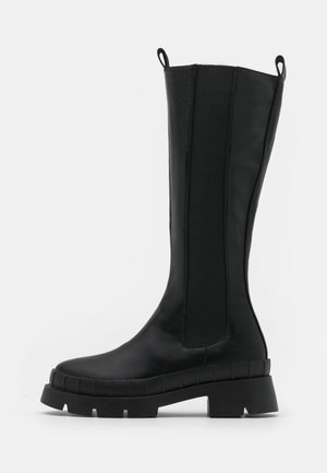 Bottes noires montant jusqu'au genou en cuir avec des panneaux latéraux élastiques et une semelle épaisse en caoutchouc texturée. Équipées de languettes pour un enfilage facile.