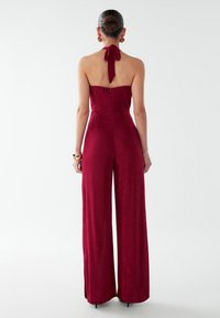 Mono burdeos de cuello halter con corsé ajustado, silueta de pierna ancha y cremallera en la espalda. Tejido suave y texturizado con detalle de lazo en el cuello.