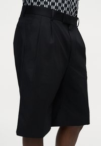 Shorts noirs sur mesure longueur genou avec des plis et des passants de ceinture, portés avec une chemise noire et blanche à motifs.