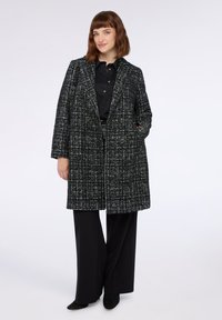 Cappotto lungo a scacchi bianco e nero con tessuto testurizzato, bottoni doppi, e ampi revers, abbinato a pantaloni neri a gamba larga.