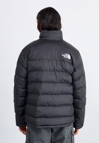 Zwarte geïsoleerde pufferjack met een hoge kraag, voorzien van horizontale stiksels en een logo op de linkerschouder. Glad, glanzend materiaal.