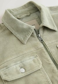 Veste en velours côtelé vert clair avec fermeture éclair, col pointu et poches avant à rabat boutonné.