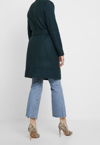 Person iført en mørkegrøn bæltekappe, lyseblå cropped jeans og højhælede sko med slangeskinds mønster, stående mod en ensfarvet baggrund.