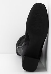Botín negro con parte superior de ante, suela de goma gruesa con patrón texturizado, detalle de cremallera lateral y diseño de punta ligeramente afilada.
