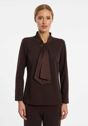 Femme portant un haut marron foncé à manches longues avec un grand nœud au niveau du cou, debout devant un fond blanc uni.