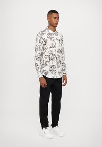 Camisa blanca de botones con un patrón floral negro, de mangas largas y con cuello estándar, combinada con pantalones negros y zapatillas blancas.