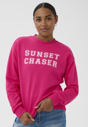 Femme aux cheveux foncés vêtue d'un sweat-shirt rose vif portant l'inscription "SUNSET CHASER", tenant l'ourlet, et portant un jean bleu sur un fond gris uni.