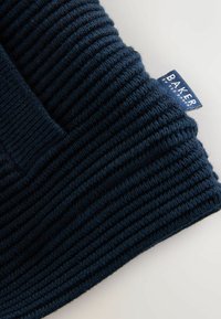 Marineblauwe gebreide beanie met ribbeltextuur en een label met de tekst "BAKER." De stof lijkt zacht en rekbaar.