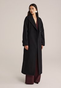 Langer schwarzer Mantel mit maßgeschneidertem Design, breitem Revers, zwei Knöpfen und Seitentaschen, getragen über einem bordeauxfarbenen Outfit.