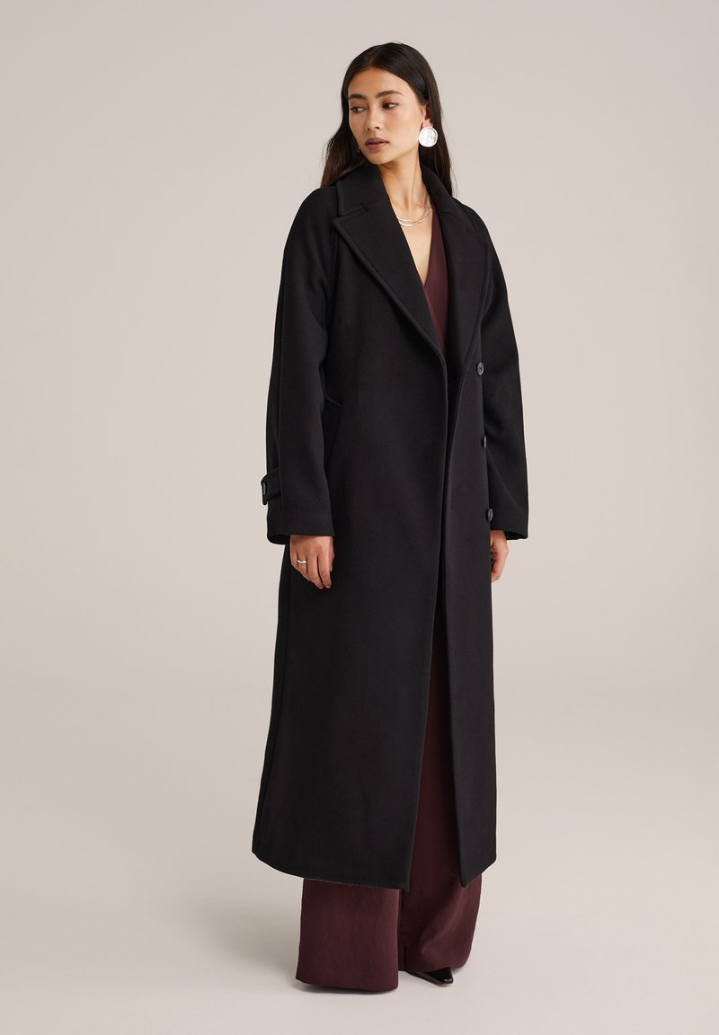 Langer schwarzer Mantel mit maßgeschneidertem Design, breitem Revers, zwei Knöpfen und Seitentaschen, getragen über einem bordeauxfarbenen Outfit.