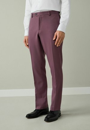 Férfi, aki testhezálló, szilvakék színű nadrágot, fehér inget és fekete alkalmi cipőt visel, és sima háttér előtt áll.