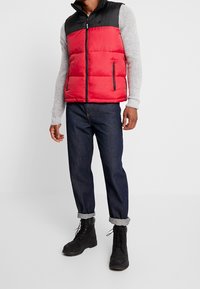 Rote und schwarze Daunenweste mit Reißverschluss vorne und Seitentaschen, kombiniert mit dunkelblauen Jeans mit umgeschlagenen Säumen und schwarzen Schnürstiefeln.